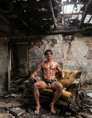 A muscular, semi-nude man poses seated in an old armchair with yellow upholstery, his torso shiny as if wet or oiled, defined abs and dark shorts, inside an abandoned and burned room. The space has peeling walls, damp stains, debris on the floor, broken furniture, and a partially destroyed ceiling with charred beams and openings through which light enters. The scene combines a decadent urban aesthetic, industrial ruin, and intense physical portraiture, with a raw and dramatic atmosphere marked by the contrast between the athletic body, the dirtiness of the surroundings, and the destruction of the building.