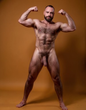 hombre culturista de 35 años posando desnudo de cuerpo completo, sonriente, tensa los bíceps, tiene un pene de 40 cm de longitud y 7 cm de diámetro, grandes dorsales, piernas, cintura estrecha, un poco calvo, grandes pezones rosas. Tiene un pene de 40 cm de longitud y 7 cm de diámetro. Tiene mucho vello en pecho y brazos y piernas. Tiene testículos de 10 kg de peso y están colgando. Tiene pies con sus uñas rojas. Fondo color amarando. Tiene el pene empalmado y eyaculando. 