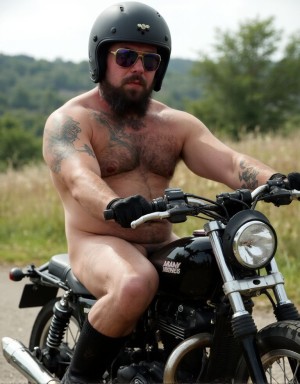 Motorista musculoso, tipo fofisano, con barba larga y tatuajes, desnudo en su foto mostrando el pene erecto visible, lleva casco negro y botas puestas, se halla en un paraje natural subido en su moto. 