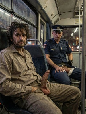 Un hombre desaliñado y cubierto de suciedad está sentado en el interior de un autobús urbano, con el pelo revuelto, barba descuidada y una expresión cansada y tensa mirando hacia delante. Viste una camisa beige arrugada y pantalones manchados, ambos muy desgastados, y sostiene una mano vendada o envuelta con una tela marrón que destaca sobre su regazo. A su lado, en el asiento contiguo, aparece un agente uniformado con gorra, placa y cinturón de servicio, sentado con postura seria y vigilante. El ambiente nocturno se percibe a través de las ventanas mojadas, con luces borrosas de la calle, asientos oscuros, barras metálicas y el interior estrecho y deteriorado del transporte público creando una escena urbana sombría, inquietante y cargada de tensión.