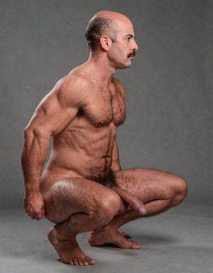 Nude man posing in a squat against a gray studio background, seen in profile with his torso upright, showing a muscular build, defined abs, strong arms, and abundant body hair on his chest, abdomen, pubic area, and legs. He has a partially bald head, a dark mustache, and a serious expression while looking to one side. The low pose keeps his feet on the floor and his hands beside his hips, highlighting defined thighs, calves, and shoulders. The penis is visible between the legs, in a sober artistic male nude composition with even lighting and a classic photographic aesthetic.