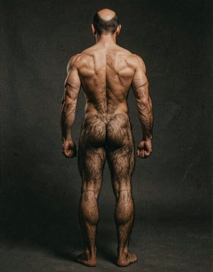 Mike Morris, a 32-year-old extremely lean Caucasian male at only 8% body fat, stands completely naked in a strict military attention pose seen from directly behind in a full posterior view. His back is perfectly straight, shoulders pulled back, arms rigid at his sides with fists clenched, legs straight and together, head held high. The shot captures him from the heels of his feet all the way up to the top of his bald head. His advanced Norwood III-IV baldness is clearly visible from behind, with a smooth tanned scalp on the crown and short dense dark brown hair only on the sides and lower back of the head. His back is highly defined and vascular: wide lats tapering dramatically to a very narrow, tight waist, thick trapezius muscles, and dense dark brown body hair lightly covering the shoulders and becoming much thicker and denser across the lower back and lumbar area, especially between the lats. His ass is tight, muscular and round with almost no fat, the glutes clearly separated. Thick dark hair continues down onto his powerful diamond-shaped calves and the backs of his slightly hairy feet. Dramatic directional studio lighting from above and slightly to the side creates strong highlights on his dry, shredded skin and deep shadows that accentuate every muscle fiber, the texture of his dense back hair, and the extreme vascularity and definition of his 8% body fat physique. Photorealistic, high-contrast, masculine 1970s Colt Studio aesthetic, grainy film texture, commanding and powerful presence. Las piernas están separadas 80 cm.  Se aprecia el surco entre glúteos respingones  muy abierto con una separación de 4 centímetros entre glúteos. 