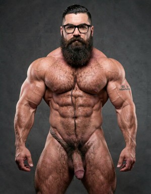 A highly detailed full-body nude photograph of a 40-year-old dominant muscle bear named Hipsterbarba, standing confidently in a well-lit studio with dramatic lighting that accentuates every muscle. He is 180cm tall, 80kg with 8% body fat, extremely muscular and symmetrical. Massive rounded hairy pecs with clear separation and dark pointed nipples, shredded six-pack abs with deep cuts and visible obliques forming a strong V-taper, wide shoulders, vascular arms, huge quads and defined legs. Very dense black masculine body hair covering his chest, running down in a thick treasure trail to a very bushy dark pubic area. He has a perfectly groomed long Bandholz beard that reaches mid-chest, thick mustache with curled upward spiral ends, modern high fade undercut hairstyle, black hipster glasses, and a small tattoo on his right bicep that reads 