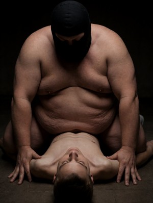 Un hombre corpulento y desnudo de torso, con pasamontañas negro que oculta casi todo su rostro, está arrodillado sobre otro hombre tumbado boca arriba en el suelo, sujetando la postura con las manos apoyadas a ambos lados del cuerpo del que yace debajo. La escena tiene una iluminación oscura y dramática, con fondo negro y ambiente cerrado, resaltando los hombros, brazos, abdomen y pliegues de piel del hombre enmascarado, así como el cuello extendido, el pecho descubierto y el rostro relajado del hombre tendido. La composición transmite tensión física, dominación, vulnerabilidad y una atmósfera íntima de estética cruda, corporal y teatral.