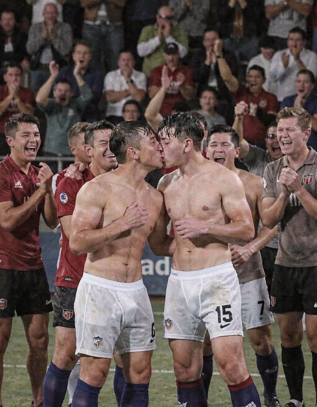 Two shirtless footballers kiss in the center of the field during an intense celebration, surrounded by teammates who applaud, laugh, and shout euphorically in front of stands full of fans. Both wear white uniform shorts, dark socks, and have sweaty torsos, with visible mud or moisture after the effort of the match; one of them wears the number 15. Around them, several players in maroon jerseys with the team crest celebrate the victory or a decisive moment, creating a football scene charged with emotion, camaraderie, collective joy, and a nighttime stadium atmosphere.