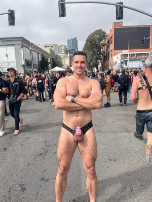 Un hombre posa en mitad de una calle urbana cerrada al tráfico durante un evento multitudinario, con los brazos cruzados y una expresión sonriente y segura. Lleva únicamente un arnés tipo suspensorio negro con tachuelas y una pieza frontal rosa que cubre el pene, dejando al descubierto el torso, las piernas y gran parte de las caderas. Al fondo hay una multitud diversa con ropa de fiesta, cuero y prendas reveladoras, además de semáforos, edificios, árboles y carpas blancas, creando un ambiente de celebración callejera tipo festival o desfile del orgullo. La escena transmite una estética queer, festiva y exhibicionista, con luz diurna, cielo nublado y una atmósfera abierta y concurrida.