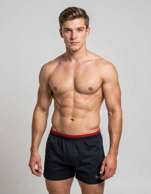 Hombre joven de complexión atlética posando de pie frente a un fondo gris claro de estudio, con el torso desnudo y una musculatura marcada en pecho, abdomen, hombros y brazos. Lleva pantalón corto deportivo negro con cintura elástica roja, dejando visibles las piernas, y mantiene una postura frontal relajada con los brazos a ambos lados del cuerpo. Su expresión es seria y neutra, mirando directamente a la cámara, con el pelo castaño corto peinado hacia un lado. La iluminación uniforme resalta la definición muscular, el aspecto fitness y el estilo de retrato deportivo minimalista.