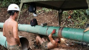 Two workers are working outdoors in a construction area surrounded by dirt, vegetation, and large-diameter green industrial pipes. One of them, shirtless, wearing a white helmet and work pants stained with mud, stands watching the intervention on the pipeline. The other is lying on his back under the pipe, also shirtless and wearing red gloves, using a power tool that generates intense sparks on a welded joint or metal repair. Around them there is work equipment, a hanging cap, an improvised shade screen, and tools resting on the ground, creating a scene of maintenance, welding, and pipe repair in rustic conditions.