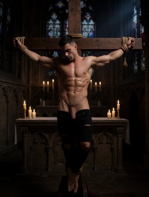 Un hombre musculoso y semidesnudo aparece atado con cuerdas a una gran cruz de madera dentro de una iglesia oscura, con los brazos extendidos y la cabeza inclinada en una pose dramática y penitente. Su torso definido queda iluminado por la luz tenue de varias velas sobre el altar, resaltando abdominales, pecho y brazos, mientras lleva unos vaqueros negros rotos y bajados lo suficiente para dejar visible el pene flácido. El entorno gótico, con vidrieras al fondo, columnas sombrías y piedra tallada, crea una atmósfera religiosa, intensa y teatral, mezclando crucifixión, desnudez masculina, bondage y estética sacra.