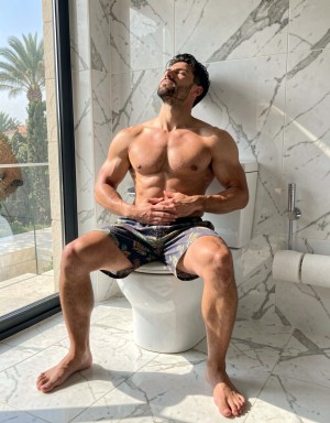 Un hombre musculoso y sin camiseta está sentado sobre un inodoro blanco en un baño moderno revestido de mármol blanco con vetas grises, iluminado por una luz solar intensa que entra por un gran ventanal lateral. Lleva bañador corto estampado, las piernas abiertas y los pies descalzos apoyados en el suelo de mármol, mientras mantiene las manos entrelazadas sobre el abdomen y la cabeza inclinada hacia atrás con los ojos cerrados, en una postura relajada y contemplativa. A la derecha aparece un portarrollos con papel higiénico y una ducha higiénica metálica, y al otro lado del cristal se distinguen palmeras, una terraza y vegetación exterior, aportando una sensación de lujo, intimidad y calor mediterráneo.