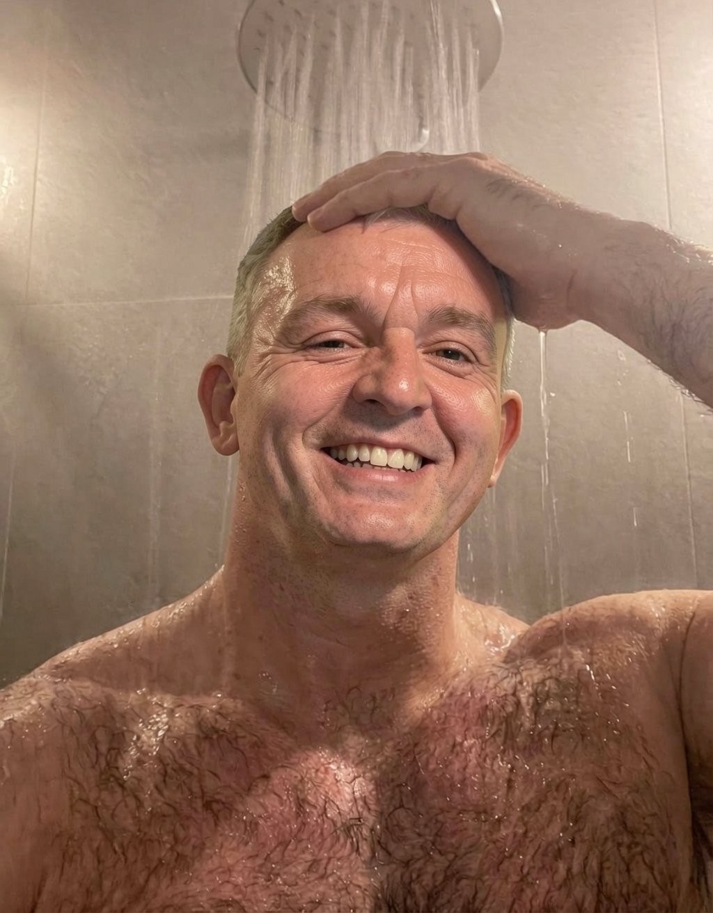 Un hombre sonriente de mediana edad aparece bajo la ducha, con el agua cayendo directamente sobre su cabeza desde un rociador redondo situado arriba. Está sin camiseta, con el torso mojado y vello en el pecho, mientras una mano descansa sobre su cabeza en un gesto relajado y espontáneo. La escena transmite cercanía e informalidad, como un selfi en el baño durante una ducha, con azulejos beige de fondo, vapor y gotas de agua visibles sobre la piel. Su expresión alegre, la iluminación cálida y el encuadre cerrado resaltan el rostro, la humedad del ambiente y la sensación de bienestar.