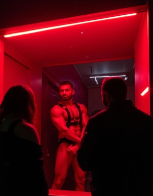 en un escaparate con luz roja un hombre musculoso vestido con suspensorio y arnés espera clientela, los clientes miran desde fuera desde la calle