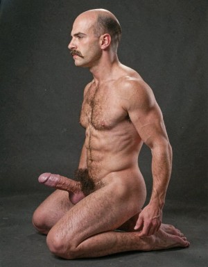 Hombre desnudo arrodillado de perfil sobre un fondo gris oscuro de estudio, con las piernas dobladas bajo el cuerpo y la espalda erguida en una postura rígida y posada. Tiene la cabeza calva, bigote espeso, rostro serio mirando hacia la izquierda, torso musculoso con pecho velludo, abdomen marcado y brazos definidos. La iluminación resalta el brillo de la piel, los hombros y la musculatura. El vello púbico es visible y el pene aparece erecto hacia delante, destacando en la composición frontal del cuerpo. La escena tiene un estilo fotográfico sobrio, minimalista y de retrato anatómico masculino.