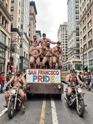 A group of men with beards, sunglasses, and leather harnesses takes part in a Pride parade on an urban avenue surrounded by skyscrapers, riding on a wooden float with the sign “SAN FRANCISCO PRIDE” painted in large colorful letters. They are almost naked, wearing bondage-style straps, boots, and leather-aesthetic accessories, posing festively and confidently while two also semi-naked motorcyclists escort the platform on both sides. The scene conveys an atmosphere of LGBTQ+ celebration, queer pride, community, street parade, and leather culture in the heart of the city, with spectators watching from the sidewalks and a bus in the background.