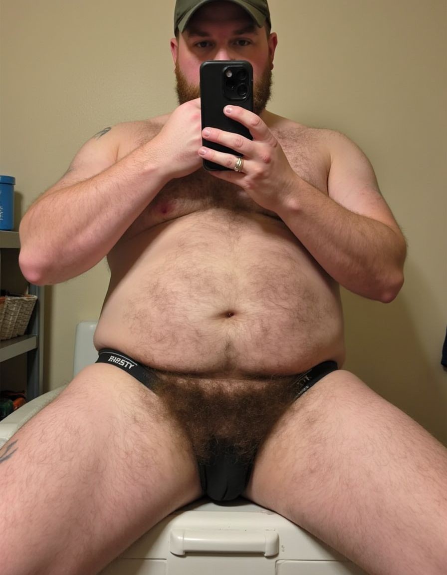Stocky bearded man sitting in front of a mirror, taking a selfie with a phone that covers part of his face. He wears a dark cap and a tight black jockstrap with an elastic waistband, leaving visible his bare torso, hairy chest and abdomen, sturdy arms, and open legs with hairy thighs. His bulge is contained by the front of the garment, with no explicit nudity. The scene looks like a simple bathroom or room, with a beige wall, a white seat, a side shelf with objects, and warm interior light that highlights an intimate, casual composition.