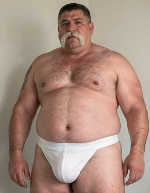 Un hombre de sesenta años, con una constitución fuerte y robusta, pesando 140 kilos, presenta una barriga impresionante y prominente, con un contorno redondeado que se extiende por debajo de su cintura. Su rostro está adornado con un bigote estilo Dalí, grueso y bien definido, de color blanco, que le confiere un aire de distinción y elegancia. Viste slips blancos ajustados que marcan claramente la silueta de su pene, que aunque es pequeño, tiene un grosor notable y se erige con confianza. La ausencia de camiseta permite apreciar su torso peludo, con un vello abundante y blanco que cubre su pecho y abdomen, resaltando su masculinidad y edad madura. Su piel es suave y arrugada, con una textura que refleja su edad y experiencia. La imagen debería ser hiperrealista, con detalles de alta calidad, capturando la textura de su piel, el brillo de su bigote blanco y la suavidad de su vello corporal. La iluminación debería ser suave, con sombras que realcen la forma de su cuerpo y resalten su presencia imponente, destacando su peso y su volumen. El fondo debería ser neutro, para que el hombre sea el centro de atención, y su presencia llene todo el espacio. El estilo de la imagen debería ser fotográfico, con un enfoque en la realidad y la autenticidad, sin elementos distractores o artificiales. La imagen debería transmitir una sensación de confianza y seguridad, como si el hombre estuviera disfrutando de su propia compañía y de su cuerpo maduro y fuerte.