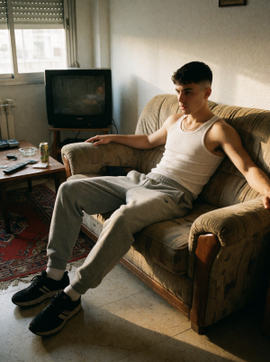 Un hombre joven está sentado de forma relajada en un sofá beige desgastado, con los brazos extendidos sobre los reposabrazos y la mirada seria dirigida hacia un lado. Lleva una camiseta blanca de tirantes, pantalones grises de chándal y zapatillas negras, iluminado por una luz cálida que entra desde la ventana y proyecta sombras sobre el suelo. La estancia tiene un aire doméstico y algo antiguo, con una televisión de tubo sobre un mueble, una mesa baja con mandos, cenicero y una lata, una alfombra roja estampada y paredes claras, creando una escena íntima de salón cotidiano con ambiente tranquilo y melancólico.