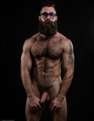 Fotografía hiperrealista de un hombre adulto musculoso pero muy delgado a la vista desde las rodillas para arriba hasta la cabeza, cuerpo muy delgado pero con musculación marcada y definida, rostro masculino idéntico a la imagen de referencia totalmente desnudo con polla erecta de treinta centímetros de longitud y ocho centímetros de diámetro, tiene rasgos marcados duros expresivos, barba densa poblada tupida oscura estilo hipster cubriendo completamente mandíbula mentón mejillas con grosor uniforme textura abundante muy definida, bigote denso tupido oscuro conectado perfectamente con la barba completa cubriendo completamente el labio superior, pelo oscuro medio peinado completamente recto hacia arriba con volumen considerable estilo hipster moderno quiff puntas verticales texturizadas hacia el cielo con altura prominente, cejas gruesas oscuras pobladas muy marcadas definidas, gafas de montura completa negra cuadradas grandes estilo hipster moderno sobre el puente de la nariz enmarcando unos ojos oscuros intensos que miran directamente a cámara con expresión seria penetrante confiada, expresión facial seria masculina sin sonrisa con mirada directa desafiante, pómulos marcados definidos angulares, nariz recta proporcionada, labios medios apenas visibles completamente cubiertos por bigote denso que conecta con la barba hipster completa, torso desnudo muy musculoso con pectorales enormes desarrollados y definidos y velludos peludos, hombros anchos musculosos deltoide prominentes, brazos musculosos gruesos con bíceps y tríceps marcados visibles a ambos lados del cuerpo, abdomen tonificado marcado visible desde la cintura, piel con textura realista detallada poros imperfecciones sutiles, vello corporal denso oscuro abundante cubriendo pectorales abdomen brazos hombros con textura natural masculina muy visible, tatuaje en tinta negra en el bíceps del brazo derecho con la palabra exacta (("hipsterbarba")) en tipografía negra sans-serif clara legible tatuada sobre la piel del brazo de tamaño mediano visible proporcionado, iluminación dramática lateral direccional que crea contraste alto marcado entre luces brillantes y sombras profundas resaltando la estructura facial angular la barba hipster densa las gafas cuadradas grandes el vello corporal abundante la musculatura del torso los brazos y el tatuaje con la palabra (("hipsterbarba")) en el bíceps derecho, fondo completamente oscuro negro liso uniforme desenfocado sin distracciones, fotografía de alto contraste con grano fotográfico sutil visible estilo retrato hipster masculino fitness, enfoque nítido absoluto perfecto en el rostro especialmente en los ojos detrás de las gafas la barba hipster densa el bigote tupido el pelo vertical el vello corporal el torso musculoso y el tatuaje con la palabra "hipsterbarba" a la vista en el brazo derecho, encuadre vertical desde la cintura hacia arriba mostrando completamente todo el torso superior los brazos musculosos y la cabeza, calidad fotográfica hiperrealista 8K máxima definición detalle extremo, renderizado cinematográfico fotorrealista profesional. Está totalmente desnudo.