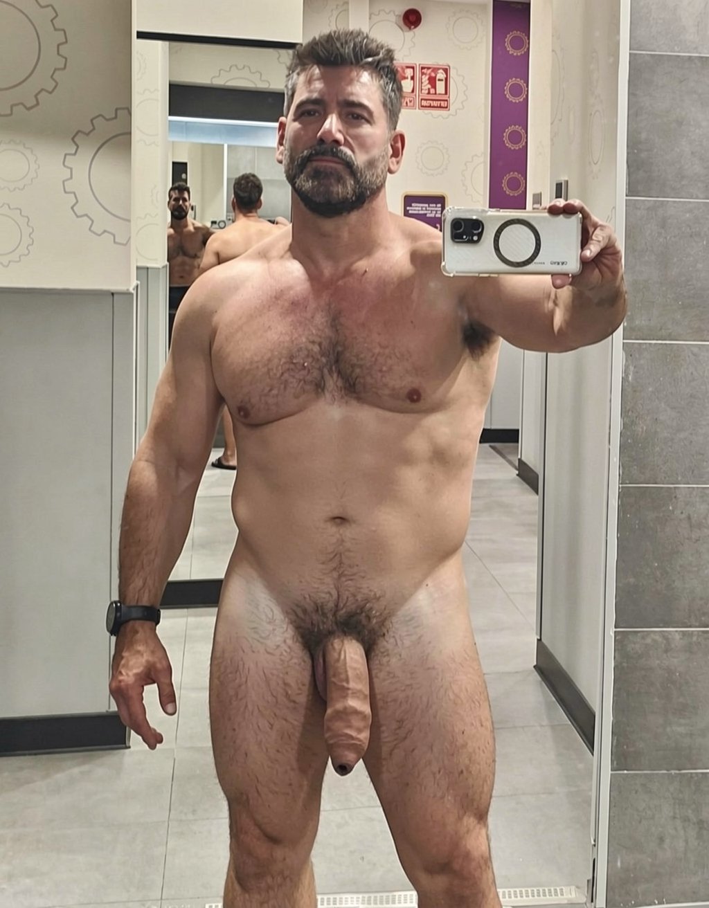 Naked man posing in front of a mirror in a gym locker room or bathroom, holding a white phone in one hand to take a selfie. He has a muscular and robust build, a broad hairy chest, a full beard, short dark hair, and a serious expression. The body is shown from the front, with strong arms, a naturally defined abdomen, visible pubic hair, and a flaccid penis hanging between the legs. He wears a black bracelet or watch on his left wrist. In the background, other mirrors, light-colored walls with gear drawings, gray tiles, tiled flooring, and emergency signage are reflected, creating a modern and bright locker-room atmosphere.