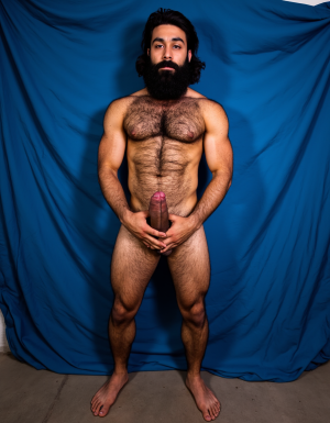 Enomao. Hombre de 35 años, cuerpo completo de pies a cabeza, pose frontal de pie, cuerpo delgado, rostro rudo con mandíbula cuadrada y ceño fruncido, barba abundante negra descuidada de varios días, nariz ancha, cicatriz pequeña en la ceja izquierda, mirada intimidante, pelo negro largo y despeinado, cuello grueso con tendones marcados, hombros anchos y trapecios abultados, pectorales enormes y duros con sombras profundas bajo cada músculo, pezones oscuros rodeados de vello negro denso, vello negro abundante cubriendo todo el pecho extendiéndose hacia el abdomen, abdominales definidos con seis bloques marcados y venas visibles, línea de vello negro desde el ombligo bajando, oblicuos duros con sombras que acentúan cada fibra muscular, brazos con bíceps y tríceps marcados y muy definidos, venas prominentes en los antebrazos, vello negro espeso en antebrazos y dorso de las manos, manos con nudillos anchos y dedos fuertes, piel con suciedad natural como grasa de trabajo y tierra en pecho brazos y piernas, manchas de mugre en los antebrazos y marcas de sudor seco, muslos marcados con cuádriceps separados y definidos cubiertos de vello negro abundante, pantorrillas duras en tensión con vello oscuro, pies descalzos plantados en suelo de hormigón, iluminación lateral dura que proyecta sombras profundas en cada grupo muscular resaltando la masa y la fuerza bruta, ambiente de atmósfera cruda y masculina, fotografía hiperrealista, textura de piel visible con poros y imperfecciones. Tiene un pene erecto masculino de 38,5 cm de longitud y 8,5 cm de diámetro, proporciones perfectas, glande grueso y pronunciado con corona marcada, color rosado oscuro en la cabeza contrastando con el tono bronceado del tronco, venas gruesas y ramificadas recorriendo toda la extensión del falo desde la base hasta el glande, piel tensa y brillante por la erección máxima, prepucio retraído mostrando el frenillo, testículos grandes y pesados colgando en escroto arrugado, cubiertos de vello púbico oscuro y denso, pelo rizado extendiéndose hacia la base del pene, piel escrotal con textura visible y poros definidos, venas visibles en los testículos, posición frontal ligeramente angular, iluminación de estudio con sombras suaves que resaltan cada relieve y textura, fotografía hiperrealista, ultra alta definición 4K, texturas extremadamente detalladas, colores naturales de piel humana, acabado cinematográfico.

El fondo de la imagen es un azul ultramar intenso y profundo, con ondas de tela que ondean como si fueran arrastradas por el viento, creando un movimiento dinámico con la figura. La textura ondulante, similar a la seda, añade una capa de profundidad y sensibilidad a la escena, contrastando con la crudeza y pasión del acto sexual.

La fotografía realista, con genitales anatómicamente correctos y proporciones naturales, sin distorsión, busca capturar la esencia cruda y visceral del momento, sin adornos ni censuras. El enfoque en el punto del pene visible desde un ángulo lateral, busca resaltar la intensidad y la conexión del hombre y su entorno, inmerso en un acto de pasión y deseo. El contenido explícito para adultos es una representación auténtica y sin tapujos de la intimidad y el deseo humano, sin rodeos ni evasivas.