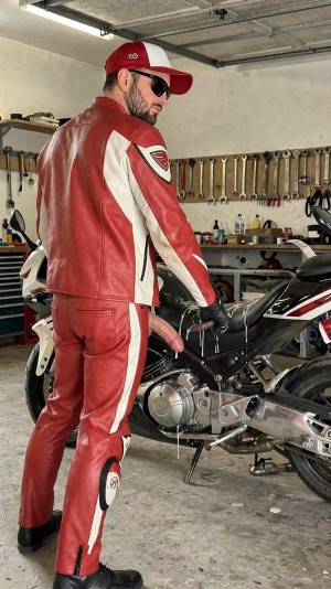 Un hombre con traje motero de cuero rojo y blanco posa de espaldas en un garaje, girando la cabeza hacia la cámara con gafas de sol oscuras, gorra roja y barba recortada. Lleva guantes negros, botas de motorista y un mono ajustado con protecciones, mientras permanece junto a una moto deportiva parcialmente desmontada o en mantenimiento, con el asiento negro y el carenado blanco, rojo y negro visibles. En la zona delantera de la entrepierna sobresale un objeto rosa con forma de pene, claramente llamativo contra el cuero rojo. Al fondo hay un banco de trabajo desordenado, herramientas colgadas en la pared, llaves fijas alineadas, cajas y piezas mecánicas, reforzando el ambiente de taller mecánico y motociclismo.