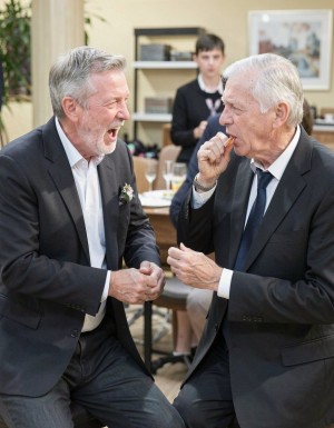 Dos hombres mayores vestidos con traje oscuro conversan animadamente en una recepción o celebración formal, sentados muy cerca frente a una mesa con copas y vajilla al fondo. El hombre de la izquierda, con pelo gris y barba recortada, lleva una flor en la solapa y aparece riendo con la boca abierta, inclinado hacia su acompañante en actitud distendida y alegre. El hombre de la derecha, de pelo blanco y corbata negra, sostiene un puro o cigarro pequeño cerca de la boca mientras escucha y gesticula con la otra mano. Detrás de ellos se aprecia un camarero desenfocado con chaleco negro, junto a estanterías, cuadros y una planta, creando un ambiente elegante de banquete, boda o evento social.