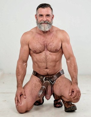 Hombre maduro y musculoso arrodillado sobre un suelo claro, con el torso desnudo, abundante vello en el pecho y abdomen marcado, mirando de frente con una ligera sonrisa. Tiene el pelo corto peinado hacia atrás y una barba gris espesa que destaca sobre el fondo blanco minimalista. Lleva un arnés de cuero oscuro ajustado a la cintura y la entrepierna, con correas y piezas metálicas que rodean el pene y dejan visibles los testículos, creando una estética fetichista de cuero. Sus manos descansan sobre los muslos, y calza sandalias negras, reforzando una pose frontal, segura y dominante.