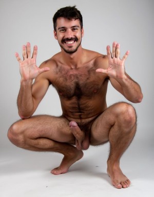 Hombre con mucho vello corporal musculoso totalmente desnudo agachado con pene erecto y saludando con la mano izquierda y rodea su pene erecto con la mano derecha. Sonríe