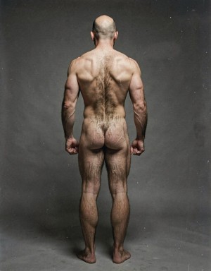 Mike Morris, a 32-year-old extremely lean Caucasian male at only 8% body fat, stands completely naked in a strict military attention pose seen from directly behind in a full posterior view. His back is perfectly straight, shoulders pulled back, arms rigid at his sides with fists clenched, legs straight and together, head held high. The shot captures him from the heels of his feet all the way up to the top of his bald head. His advanced Norwood III-IV baldness is clearly visible from behind, with a smooth tanned scalp on the crown and short dense dark brown hair only on the sides and lower back of the head. His back is highly defined and vascular: wide lats tapering dramatically to a very narrow, tight waist, thick trapezius muscles, and dense dark brown body hair lightly covering the shoulders and becoming much thicker and denser across the lower back and lumbar area, especially between the lats. His ass is tight, muscular and round with almost no fat, the glutes clearly separated. Thick dark hair continues down onto his powerful diamond-shaped calves and the backs of his slightly hairy feet. Dramatic directional studio lighting from above and slightly to the side creates strong highlights on his dry, shredded skin and deep shadows that accentuate every muscle fiber, the texture of his dense back hair, and the extreme vascularity and definition of his 8% body fat physique. Photorealistic, high-contrast, masculine 1970s Colt Studio aesthetic, grainy film texture, commanding and powerful presence.