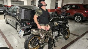 A man with a black helmet, camouflage t-shirt, tactical vest and motorcycle boots appears next to a black trail-type motorcycle in an underground parking lot, with his head tilted back and camouflage pants pulled down to his thighs, exposing the underwear area and crotch in a provocative pose. The motorcycle has rigid side cases and protection details, with the license plate visible at the rear. Around it there are several parked cars, concrete columns, tiled floor marked with parking space lines and cold lighting typical of a closed garage.