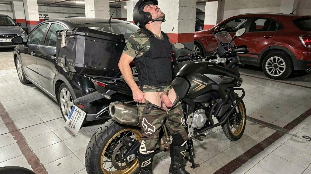 A man with a black helmet, camouflage t-shirt, tactical vest and motorcycle boots appears next to a black trail-type motorcycle in an underground parking lot, with his head tilted back and camouflage pants pulled down to his thighs, exposing the underwear area and crotch in a provocative pose. The motorcycle has rigid side cases and protection details, with the license plate visible at the rear. Around it there are several parked cars, concrete columns, tiled floor marked with parking space lines and cold lighting typical of a closed garage.