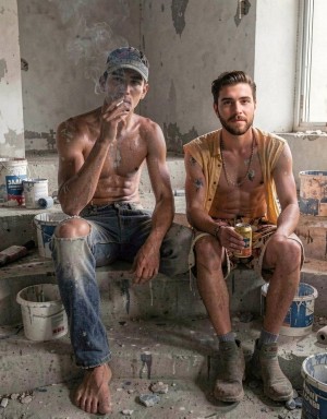 Two young men rest seated at a construction site or room under renovation, surrounded by paint buckets, brushes, rubble, and peeling walls with plaster stains. The one on the left is shirtless, covered in white paint, wearing ripped jeans, a cap, and holding a cigarette in his hand, barefoot on the dirty floor. The one on the right wears an open sleeveless yellow shirt, shorts, work boots, and holds a can, also stained with paint. The scene conveys a work break, an industrial atmosphere, home renovation, fatigue, and camaraderie between painters or bricklayers in a deteriorated space next to a window.