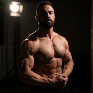 Hombre musculoso posando sin camiseta en un estudio oscuro, con el torso desnudo iluminado por una luz intensa situada al fondo a la izquierda que crea un contraste dramático de sombras sobre sus pectorales, abdominales, hombros y brazos definidos. Lleva barba recortada, pelo corto y expresión seria, mirando hacia la cámara con una postura firme y segura mientras entrelaza las manos a la altura de la cintura, resaltando las venas y la musculatura de los antebrazos. El ambiente de iluminación cinematográfica, fondo negro y estética de fitness o culturismo enfatiza la fuerza física, la definición corporal y el retrato masculino atlético.