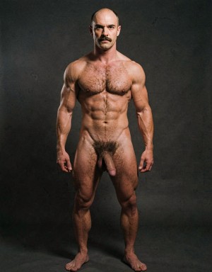 Mike Morris, a 32-year-old Caucasian male with an extremely lean and athletic physique at only 8% body fat, stands completely naked in a strict military attention pose. Full body frontal shot from the tips of his toes to the top of his head. His body is young, dry, and highly defined with sharp muscle separation, visible vascularity, and a very narrow waist. He has advanced Norwood III-IV male pattern baldness with a shiny tanned scalp on top and only short dark brown hair on the sides and back. His face is serious and commanding: square angular jaw, prominent cheekbones, thick dark handlebar mustache, heavy brows, and intense deep-set brown eyes. His massive square pectorals are pushed forward, covered in dense dark brown chest hair that forms a classic thick T-pattern running down the sternum and spreading over his sharply carved six-pack abs. Small dark nipples sit low on the pecs. A prominent trail of dark hair leads to an extremely dense, curly, dark brown pubic bush that climbs almost to his navel. His thick 14 cm flaccid cock hangs heavy over large, low-hanging, hairy testicles, the shaft skin noticeably lighter than his bronzed body. Powerful veined forearms, developed traps, defined quadriceps with clear separation, diamond-shaped calves, and slightly hairy feet complete his figure. He stands perfectly straight, chest out, shoulders back, arms rigid at his sides with fists clenched, head high and gaze fixed forward. Dramatic directional studio lighting from above and slightly to the front creates strong highlights on his dry, vascular skin and deep shadows that accentuate every muscle fiber, the texture of his dense body hair, and the extreme definition of his 8% body fat physique. Photorealistic, high-contrast, 1970s Colt Studio masculine aesthetic, grainy film texture.