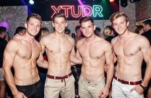 Cuatro hombres jóvenes posan sonrientes y sin camiseta en una discoteca o fiesta nocturna, mostrando torsos desnudos y físicos atléticos mientras se abrazan por los hombros y la cintura en actitud cercana y festiva. Llevan pantalones claros y oscuros con cinturones, peinados cuidados y expresión relajada ante la cámara. Al fondo hay una pista llena de gente, luces de colores, focos de ambiente y una pared de ladrillo con un luminoso de neón que dice “XTUDR”, creando una escena de ocio, música, ambiente gay-friendly y vida nocturna con estética de club urbano.