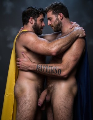 Hipsterso y Barbato son dos hombres musculosos de 30 años abrazados íntimamente, cuerpo completo visible, primer hombre llamado Hipsterso con pelo negro azulado corto despeinado hacia arriba con capa amarilla flotando detrás de su espalda, segundo hombre llamado Barbato con pelo castaño claro corto despeinado con capa azul oscura flotando detrás de su espalda, Hipsterso y Barbato ambos con rasgos faciales masculinos marcados, mandíbulas cuadradas definidas, pómulos altos, cejas pobladas oscuras, ojos marrones intensos mirándose entre ellos, narices rectas proporcionales, labios carnosos entreabiertos, expresiones de deseo intenso, torsos completamente desnudos hipermusculosos, pectorales enormes marcados y definidos cubiertos de vello oscuro denso, abdominales marcados six-pack, brazos musculosos cubiertos de vello oscuro rodeando espaldas mutuamente, venas visibles en bíceps y antebrazos, axilas con vello oscuro abundante visible, pieles bronceadas brillantes cubiertas de sudor realista con gotas visibles resbalando por pectorales y abdominales, piernas musculosas desnudas separadas cubiertas de vello oscuro denso, penes erectos enormes de 35 centímetros largos y muy gruesos con venas gruesas pulsantes marcadas recorriendo todo el tronco, glandes hinchados enormes brillantes de color rojo púrpura, testículos extremadamente grandes gordos y pesados colgando muy abajo descolgados entre las piernas con piel arrugada tensa bajo el peso,  pose abrazados estrechamente pecho contra pecho, músculos tensos, cuerpos pegados, capas flotando dramáticamente detrás de ellos, fondo oscuro difuminado dramático, iluminación cinematográfica lateral contrastada resaltando músculos sudor y eyaculación, fotografía hiperrealista homoerótica explícita, ultra detallada, 8K, máxima definición anatómica. Hipsterso y Barbato tienen cada uno un tatuaje de tinta negra sobre la piel del bíceps de sus brazos derechos en tipografía negra sans-serif de tamaño mediano claramente legible y visible y proporcionado con las siguientes letras: hipsterbarba.