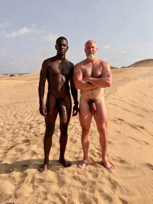 Two naked men pose standing on sand dunes in a desert landscape by the sea, under a clear and bright sky. The man on the left, with dark skin and an athletic build, looks at the camera with relaxed arms and his penis visibly hanging forward. Next to him, a man with light skin, bald and with a white beard, appears with his arms crossed over his chest, also completely naked, with his penis and testicles visible. The scene conveys an outdoor naturist portrait, with footprints in the sand, dry hills in the background and intense sunlight.