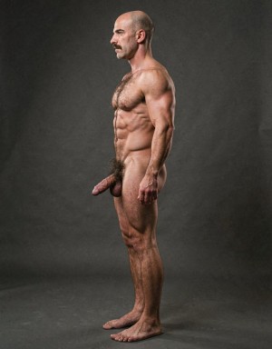 Mike Morris, a 32-year-old extremely lean Caucasian male with only 8% body fat, stands completely naked in a strict military attention pose captured in a full-body exact side profile at 90 degrees from his left side. The shot runs from the tips of his size 44 slightly hairy feet all the way up to the top of his bald head, showing his entire body without any cropping. His physique is dry, highly vascular and powerfully defined: massive square pectorals seen in perfect side silhouette, dense dark brown chest hair forming a thick T-pattern, sharply carved six-pack abs in clear profile, narrow waist, wide lats, powerful thighs and diamond-shaped calves. Dramatic directional studio lighting comes from high and slightly in front of him, creating strong rim lighting that traces the sharp outline of his entire body, the texture of his dense dark body hair on chest, lower back, ass and legs, and deep shadows that accentuate every muscle separation, vein and the extreme dryness of his 8% body fat physique. His advanced Norwood III-IV bald scalp shines under the light, with short dense dark brown hair on the sides and back of his head. His square angular face is shown in pure profile: strong jawline, prominent chin with slight central dimple, thick dark handlebar mustache, heavy brow and intense deep-set brown eye looking straight forward with a serious, commanding expression. His penis is fully visible in perfect side view, completely natural and realistic: thick, 24 cm long when fully erect, with a natural slight upward curve, prominent dorsal veins, long foreskin partially covering the pink-red glans, and realistic lighter skin tone. Below it hang his large, low-hanging testicles with wrinkled darker skin and dense dark brown hair. The extremely thick, curly dark brown pubic bush climbs almost to his navel and surrounds the base of his cock and balls generously. Every anatomical detail from his veined hands and forearms to his muscular ass, hamstrings, diamond calves and hairy toes is perfectly proportioned and free of any deformation. Photorealistic, high-contrast, masculine 1970s Colt Studio aesthetic with subtle grainy film texture.