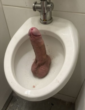 Dildos. Imagen surrealista, alguien ha colocado un consolador (dildo) de color carne y muy perfecto en un urinario público, un chico hetero lo mira con asco. 