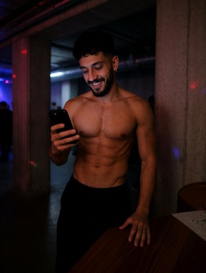 Hombre joven sin camiseta apoyado junto a una columna de hormigón en un espacio interior oscuro con ambiente de club o local nocturno, iluminado por luces tenues azuladas y pequeños reflejos rojos. Sonríe mientras mira la pantalla de un teléfono móvil negro que sostiene en una mano, con una expresión relajada y divertida. Su torso musculoso y definido, con abdominales marcados y pecho descubierto, destaca bajo la iluminación lateral, mientras lleva pantalones negros. Una mano descansa sobre una superficie de madera en primer plano, y al fondo se intuyen sombras de personas, pilares y una atmósfera urbana, íntima y nocturna.
