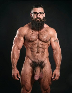 A highly detailed full-body nude photograph of a 40-year-old dominant muscle bear named Hipsterbarba, standing confidently in a well-lit studio with dramatic lighting that accentuates every muscle. He is 180cm tall, 80kg with 8% body fat, extremely muscular and symmetrical. Massive rounded hairy pecs with clear separation and dark pointed nipples, shredded six-pack abs with deep cuts and visible obliques forming a strong V-taper, wide shoulders, vascular arms, huge quads and defined legs. Very dense black masculine body hair covering his chest, running down in a thick treasure trail to a very bushy dark pubic area. He has a perfectly groomed long Bandholz beard that reaches mid-chest, thick mustache with curled upward spiral ends, modern high fade undercut hairstyle, black hipster glasses, and a small tattoo on his right bicep that reads 