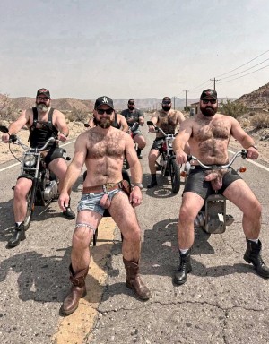 Five bearded, muscular men pose on a cracked desert road, surrounded by arid hills, scrubland, and utility poles under a clear sky. They wear biker and fetish aesthetics, with caps, sunglasses, leather harnesses, boots, and minimal clothing; several wear jockstraps or very tight shorts that leave their hairy chests exposed and explicitly outline the penis beneath the fabric. Two are seated on minibikes or small motorcycles, while another poses standing in the foreground with cowboy boots and a harness, creating a provocative biker group scene in a desert setting, abandoned road, and defiant attitude.