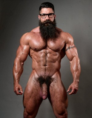 A highly detailed full-body nude photograph of a 40-year-old dominant muscle bear named Hipsterbarba, standing confidently in a well-lit studio with dramatic lighting that accentuates every muscle. He is 180cm tall, 80kg with 8% body fat, extremely muscular and symmetrical. Massive rounded hairy pecs with clear separation and dark pointed nipples, shredded six-pack abs with deep cuts and visible obliques forming a strong V-taper, wide shoulders, vascular arms, huge quads and defined legs. Very dense black masculine body hair covering his chest, running down in a thick treasure trail to a very bushy dark pubic area. He has a perfectly groomed long Bandholz beard that reaches mid-chest, thick mustache with curled upward spiral ends, modern high fade undercut hairstyle, black hipster glasses, and a small tattoo on his right bicep that reads 