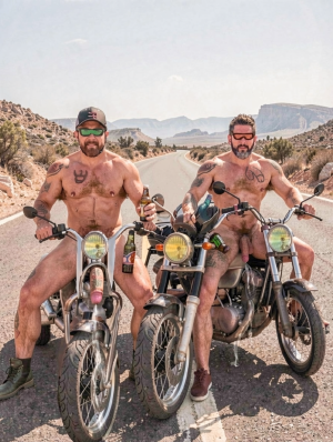 Two naked men pose seated on classic motorcycles in the middle of a desert road, surrounded by arid mountains, scrubland and a dry landscape under a clear sky. Both wear sunglasses, beards and visible tattoos on their chests and arms; the one on the left also wears a cap and holds a beer bottle while his penis is visible between his open legs on the motorcycle. The man on the right, with sports shoes, is equally naked and shows his penis while gripping the handlebars. The scene conveys a biker, rebellious and provocative aesthetic, with boots, vintage motorcycles, a lonely road and a desert route atmosphere.