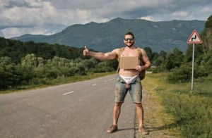Un hombre con gafas de sol está de pie en el arcén de una carretera rural, haciendo autoestop con el pulgar levantado mientras sostiene un cartón rectangular contra el pecho desnudo, como si fuera un cartel improvisado. Lleva pantalones cortos vaqueros, sandalias y una riñonera clara a la cintura, con una postura relajada y algo cómica. La carretera vacía se pierde hacia un paisaje verde de campos, árboles y montañas al fondo, bajo un cielo nublado y gris. A la derecha aparece una señal de tráfico de curva peligrosa, reforzando el ambiente de viaje, carretera secundaria, aventura y turismo rural.