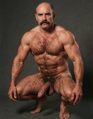 Bald, muscular man posing nude in a squat in front of a dark gray studio background, with a serious expression and looking directly at the camera. His physique is highly defined, with broad shoulders, strong arms, a hairy chest, marked abs, and sturdy legs covered in hair. He has a thick mustache that accentuates a mature and forceful appearance. The low posture, with his hands near his knees and his feet planted on the floor, leaves the penis and scrotum visible between his thighs, in a frontal, raw composition focused on male anatomy, bodily strength, and an unclothed physical portrait.