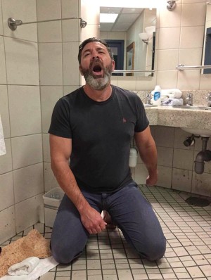 Un hombre arrodillado en el suelo de un baño público o de vestuario, vestido con camiseta negra ajustada y pantalones vaqueros azul oscuro, aparece con la cabeza ligeramente inclinada hacia atrás y la boca muy abierta, como si estuviera gritando, cantando o haciendo una expresión exagerada. Tiene barba canosa, pelo oscuro corto y una postura tensa, con una mano apoyada cerca de la entrepierna sobre el pantalón. El entorno muestra azulejos beige en las paredes, suelo de mosaico cuadrado, un lavabo de encimera con espejo, dispensadores, toallas enrolladas y tuberías visibles bajo el fregadero. En una esquina hay una papelera pequeña y una toalla o alfombrilla beige en el suelo, reforzando la sensación de baño funcional y algo descuidado.
