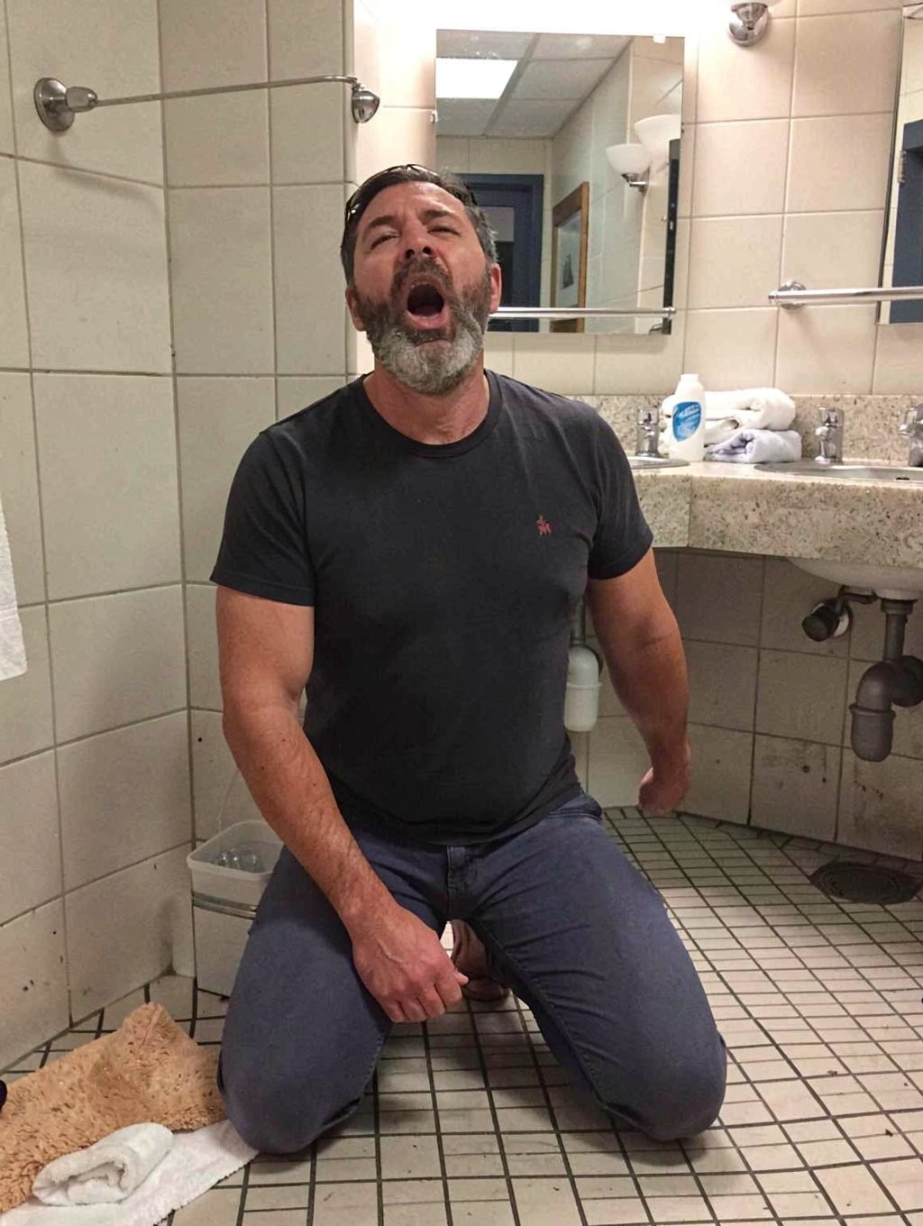 Un hombre arrodillado en el suelo de un baño público o de vestuario, vestido con camiseta negra ajustada y pantalones vaqueros azul oscuro, aparece con la cabeza ligeramente inclinada hacia atrás y la boca muy abierta, como si estuviera gritando, cantando o haciendo una expresión exagerada. Tiene barba canosa, pelo oscuro corto y una postura tensa, con una mano apoyada cerca de la entrepierna sobre el pantalón. El entorno muestra azulejos beige en las paredes, suelo de mosaico cuadrado, un lavabo de encimera con espejo, dispensadores, toallas enrolladas y tuberías visibles bajo el fregadero. En una esquina hay una papelera pequeña y una toalla o alfombrilla beige en el suelo, reforzando la sensación de baño funcional y algo descuidado.