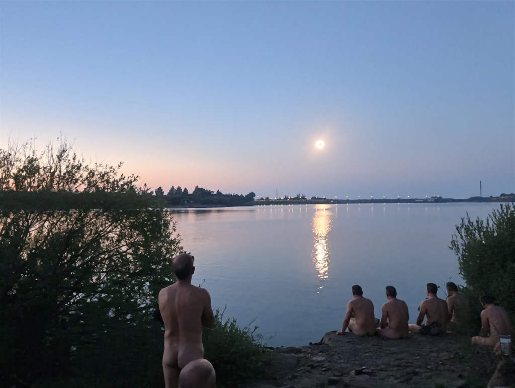 A group of naked people rests by the shore of a tranquil lake at dusk, gazing at the water and the horizon under a soft blue sky with pink tones. On the left, a standing person appears from behind among bushes, with their buttocks visible, while several seated figures on the pebbly bank look toward the bright reflection of the moon on the water's surface. The scene conveys calm and naturalness, with dark vegetation in the foreground, silhouettes of trees in the background, and a serene atmosphere of nighttime bathing, naturism, and a lakeside landscape illuminated by moonlight.