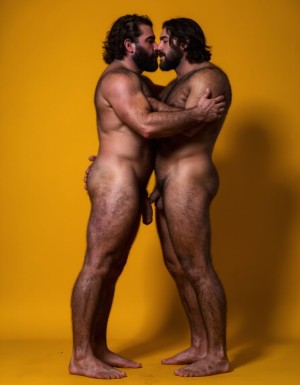 Dos guerreros musculosos delgados y altos, cuerpo completamente desnudo y velludo, mostrando polla venosa de 30 cm de longitud y 5 cm de grosor. Tienen la piel bronceada por el Sol, con pelo muy largo y barba. Tiene pies fuertes. Están contentos. Tienen músculos marcados definidos y sin nada de grasa. Tienen aspecto muy viril. Tienen cuerpo muy delgado pero con los músculos marcados y muy definidos. Están tensos haciendo fuerza para mostrar sus músculos marcados. Fondo amarillo oscuro con los reflejos de las sombras de los cuerpos. Están abrazados y besándose en la boca. 